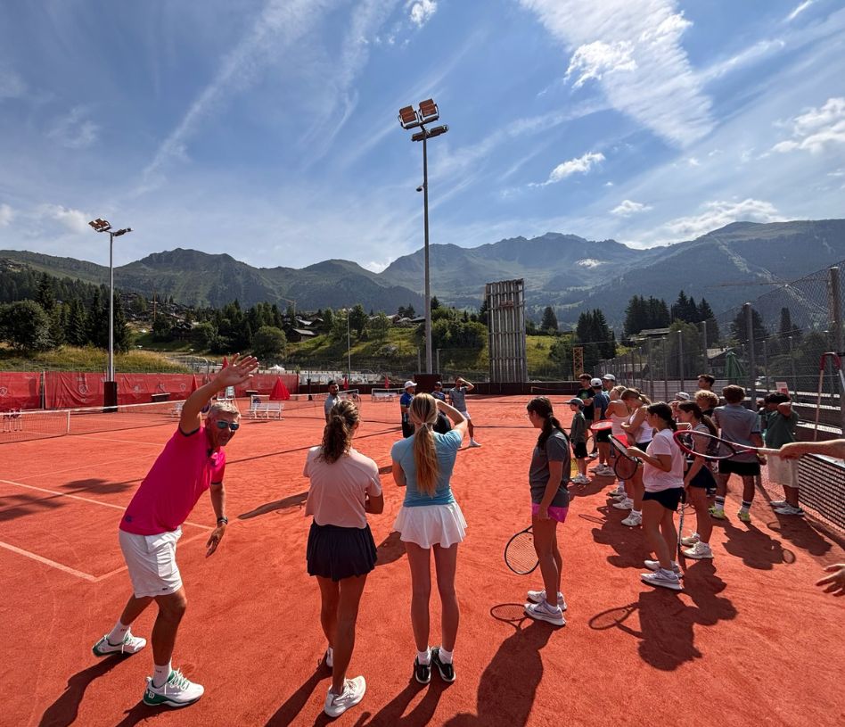 Group of junior tennis players at an academy with mountains in the background and a coach waving to the camera